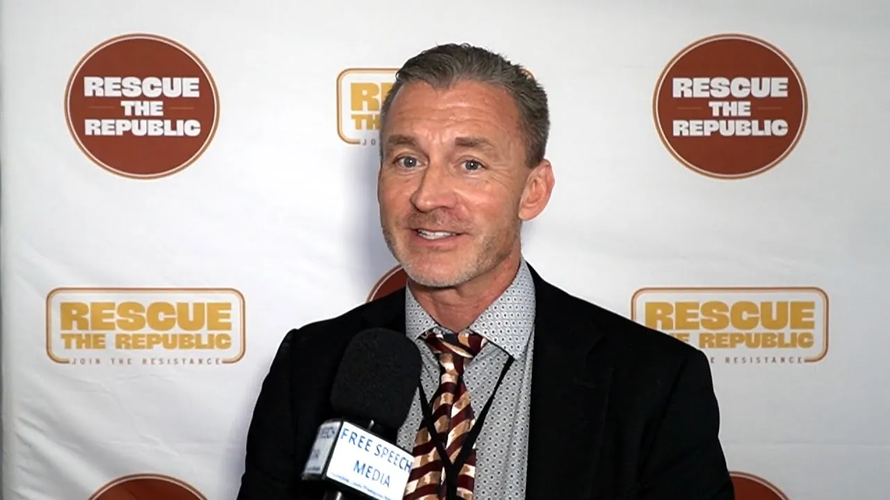 Dr Ryan Cole being interviewed in front of a Rescue the Republic backdrop, Free Speech Media mic flag in frame