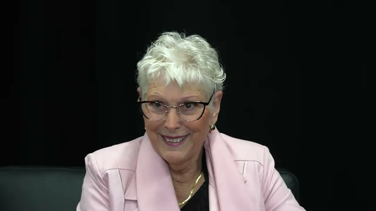 Ann Gillies seated in a pink jacket against a dark background