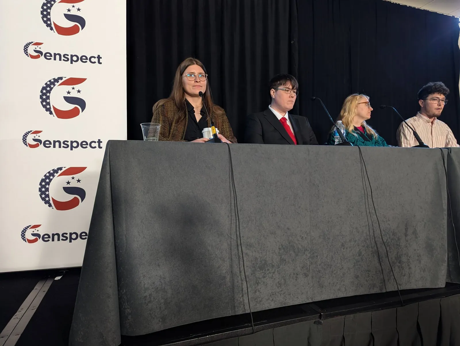A five-person panel at a draped table in front of Genspect banners
