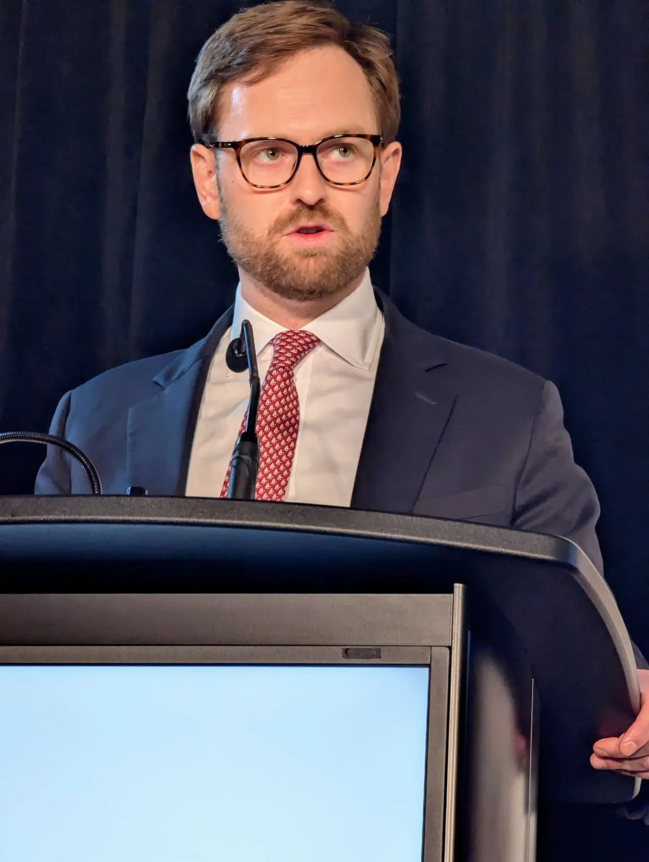 A man in glasses and a red tie speaking at a lectern