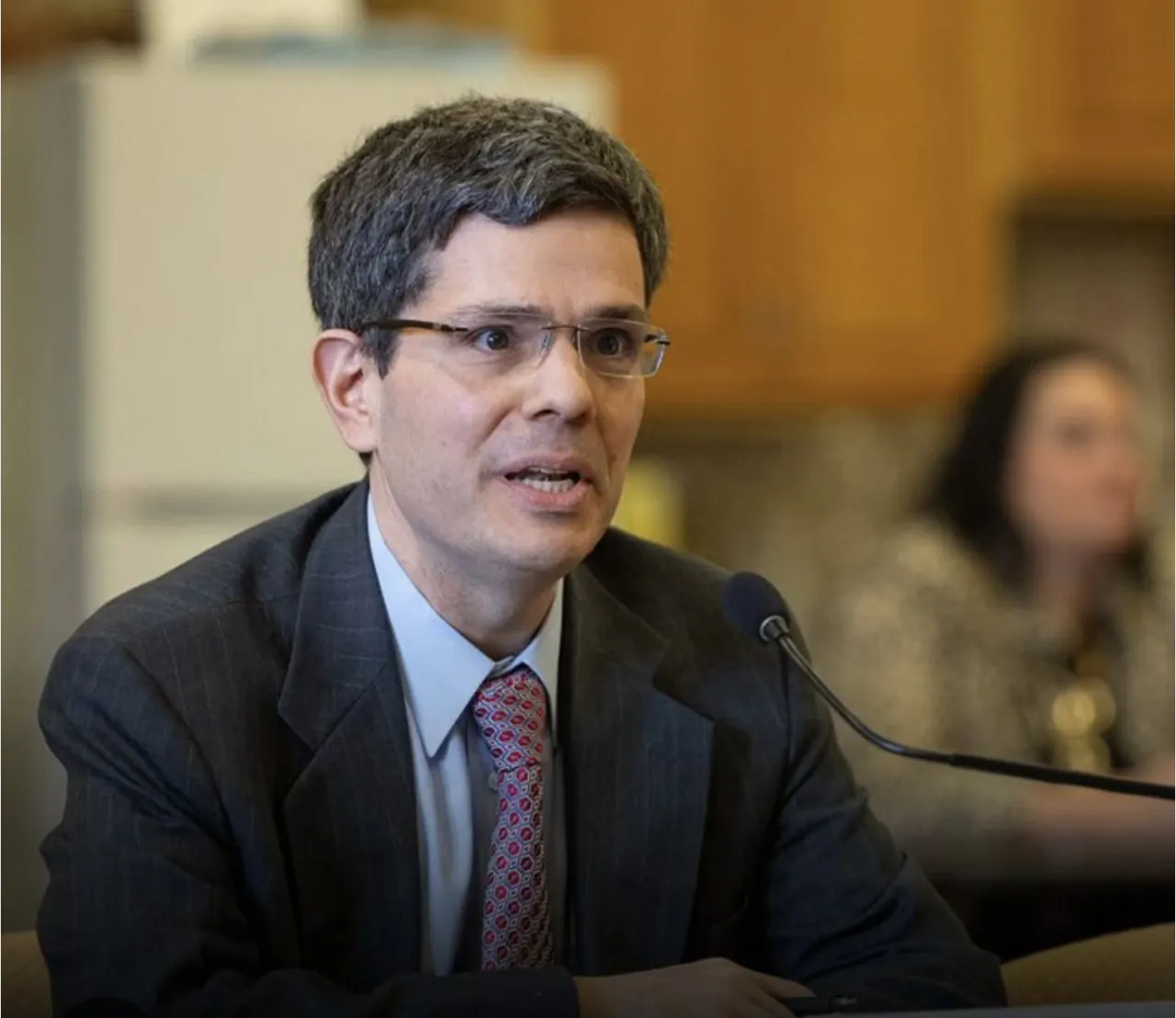 A man in a suit speaking into a microphone at a hearing table